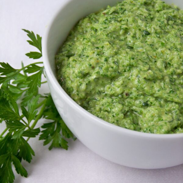 A white bowl filled with a green, textured dip or sauce sits on a white surface, with fresh parsley leaves placed beside the bowl.