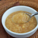 A spoonful of applesauce is held above a white bowl filled with more applesauce, placed on a woven placemat with a brown napkin underneath.