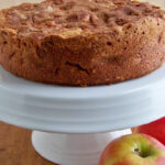 A round apple cake with a golden-brown crust sits on a white cake stand, with two fresh apples placed beside it on a wooden surface.