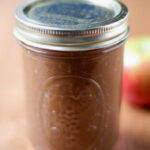 A glass mason jar filled with brown apple butter, sealed with a metal lid, sits on a wooden surface with a blurred apple in the background.