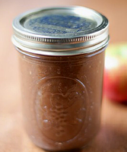 A glass mason jar filled with brown apple butter, sealed with a metal lid, sits on a wooden surface with a blurred apple in the background.