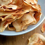 A white plate filled with thin, crispy apple chips sits on a wooden surface, with a few apple chips scattered nearby. The chips are golden brown and slightly curled at the edges.