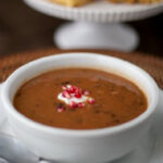 A bowl of bean soup topped with a dollop of cream and chopped red onions, served on a white plate with a spoon beside it; cornbread squares are displayed in the background on a cake stand.
