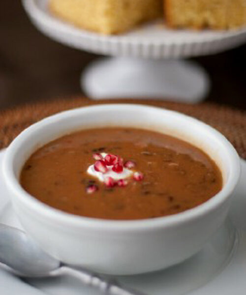 A bowl of bean soup topped with a dollop of cream and chopped red onions, served on a white plate with a spoon beside it; cornbread squares are displayed in the background on a cake stand.