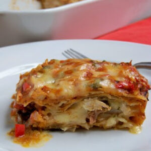 A slice of cheesy, vegetable lasagna with a golden-brown top sits on a white plate next to a fork. In the background, a baking dish contains the remaining lasagna.