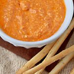 A white bowl filled with orange-red dip, likely a roasted red pepper spread, sits on a brown mat next to several long, thin breadsticks.
