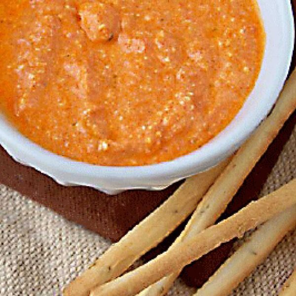 A white bowl filled with orange-red dip, likely a roasted red pepper spread, sits on a brown mat next to several long, thin breadsticks.