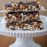 Three granola bars with chocolate chips and puffed rice are stacked on a white cake stand against a brown background.