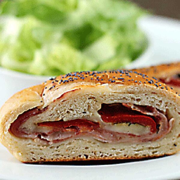 A close-up of a sliced stromboli filled with meats and cheese on a white plate, with a side of chopped green lettuce in the background.