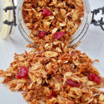A glass jar tipped over, spilling granola with oats, sliced almonds, and dried cranberries onto a white surface.