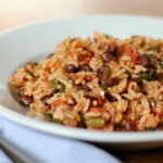 A white plate filled with a serving of rice mixed with black beans, diced tomatoes, green peppers, and herbs, sitting on a table next to a fork and a light blue napkin.