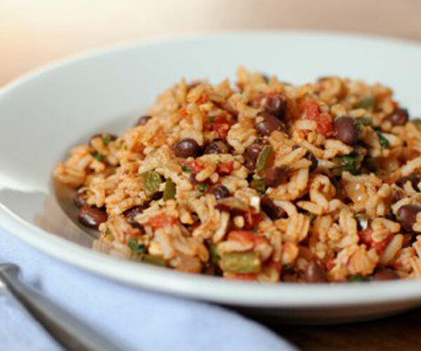 A white plate filled with a serving of rice mixed with black beans, diced tomatoes, green peppers, and herbs, sitting on a table next to a fork and a light blue napkin.