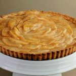 A baked pear tart with a golden crust and neatly arranged pear slices, displayed on a white cake stand against a neutral background.