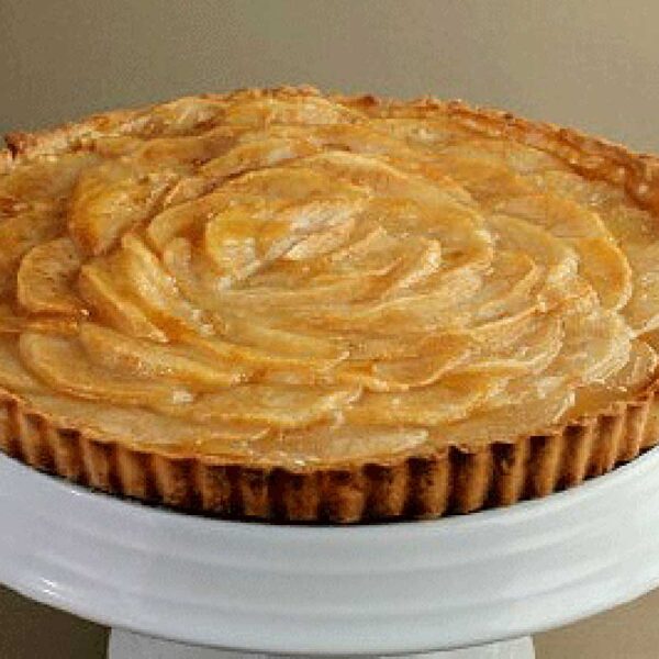 A baked pear tart with a golden crust and neatly arranged pear slices, displayed on a white cake stand against a neutral background.