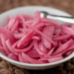 A white bowl filled with thinly sliced pickled red onions, with a metal fork resting on the bowl, placed on a woven brown mat.