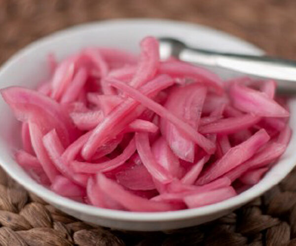 A white bowl filled with thinly sliced pickled red onions, with a metal fork resting on the bowl, placed on a woven brown mat.