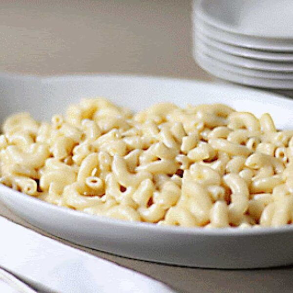 A white oval dish filled with creamy macaroni and cheese sits on a table next to a silver spoon, a white napkin, and a stack of white plates in the background.