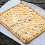 A rectangular apple tart with thinly sliced apples arranged in overlapping rows sits on parchment paper atop a wooden surface. The tart has golden-brown edges and a slightly crisp crust.