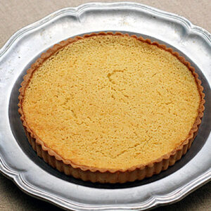 A plain baked tart crust with a golden-brown surface sits on a decorative silver plate atop a beige fabric background.