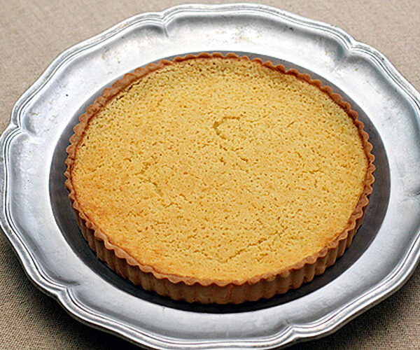 A plain baked tart crust with a golden-brown surface sits on a decorative silver plate atop a beige fabric background.