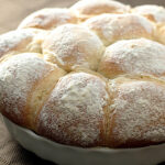 Round, golden-brown bread rolls dusted with powdered sugar, closely packed together in a white ceramic dish on a brown textured surface.