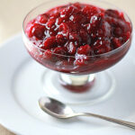 A glass dish filled with chunky cranberry sauce sits on a white plate with a silver spoon beside it.
