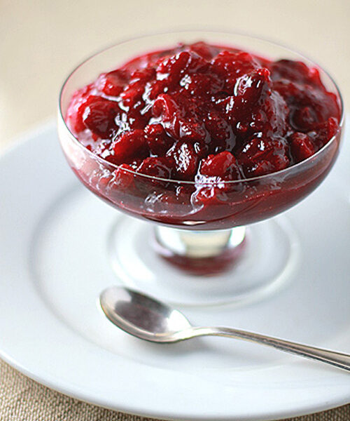 A glass dish filled with chunky cranberry sauce sits on a white plate with a silver spoon beside it.