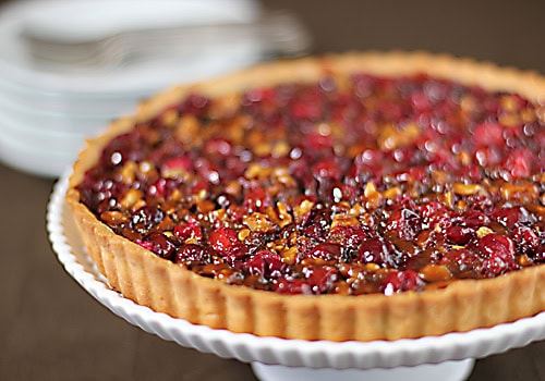 A close-up of a cranberry and nut tart with a golden crust, displayed on a white cake stand. Plates and forks are stacked in the blurred background.