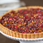 A close-up of a cranberry and nut tart with a golden crust, displayed on a white cake stand. In the blurred background, there are stacked white plates and silver forks.