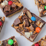Squares of chocolate dessert bars topped with assorted candy pieces, including M&Ms, nougat, and white chocolate, arranged on a white surface.