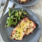 A gray plate with a mixed green salad and two slices of toasted bread topped with melted cheese and ham, garnished with chopped chives. A fork and knife rest on a white napkin nearby.