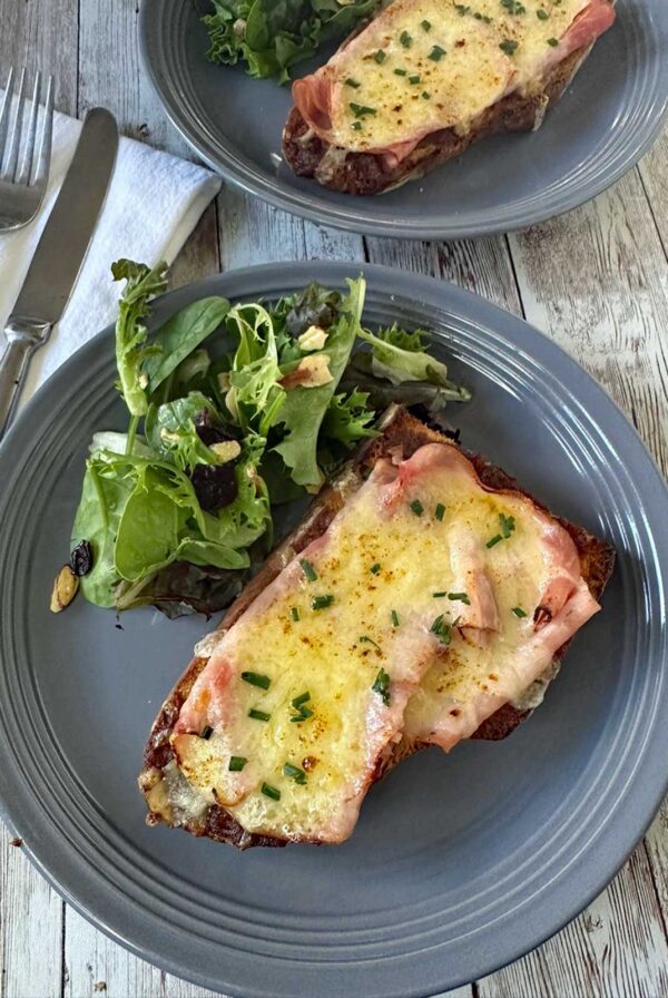A gray plate with two slices of toasted bread topped with melted cheese and ham, garnished with chopped chives, served with a side of mixed green salad. A fork and knife are next to the plate on a white napkin.