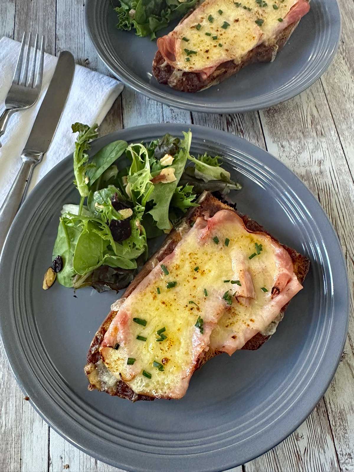 A gray plate with two slices of bread topped with melted cheese and ham, garnished with chopped chives, served alongside a small green salad. A fork and knife are placed on a napkin nearby.