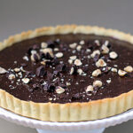 A chocolate tart with a golden crust, topped with chopped hazelnuts and pieces of dark chocolate, displayed on a white cake stand against a neutral background.