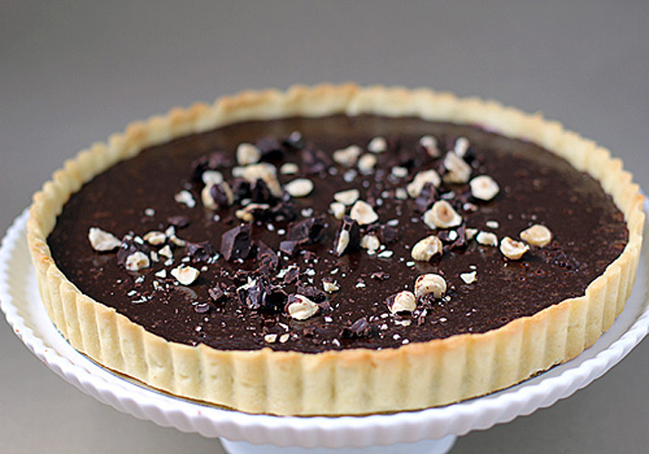 A chocolate tart with a golden crust, topped with chopped hazelnuts and pieces of dark chocolate, displayed on a white cake stand against a neutral background.
