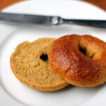 A sliced plain bagel on a white plate, with one half open-faced and the other half resting beside it. A butter knife is placed on the plate in the background.