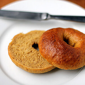 A sliced plain bagel on a white plate, with one half open-faced and the other half resting beside it. A butter knife is placed on the plate in the background.