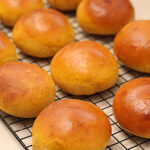 Golden brown bread rolls cooling on a black wire rack, with a light-colored countertop in the background. The rolls have a shiny, smooth surface and are arranged in rows.