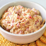 A white bowl filled with pimento cheese spread sits on a white plate, surrounded by round, golden crackers.