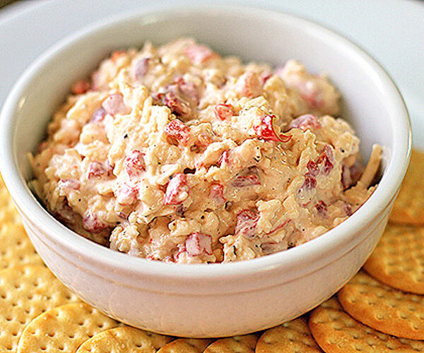 A white bowl filled with pimento cheese spread sits on a white plate, surrounded by round, golden crackers.