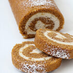 A pumpkin roll cake with cream cheese filling is dusted with powdered sugar, with two round slices cut and placed in front on a white plate.