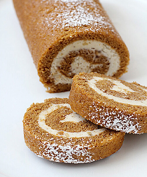 A pumpkin roll cake with cream cheese filling is dusted with powdered sugar, with two round slices cut and placed in front on a white plate.