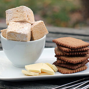 A white plate holds a stack of square cookies, several pieces of white chocolate, and a bowl filled with large, square marshmallows. The plate sits on a wooden surface outdoors.