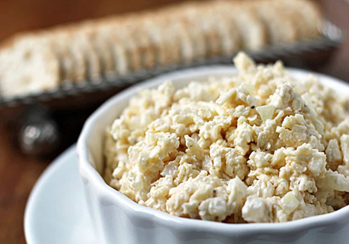A white bowl filled with creamy, chunky Sweet Onion Cheddar Spread sits on a white plate, with a row of crackers arranged on a tray in the background.