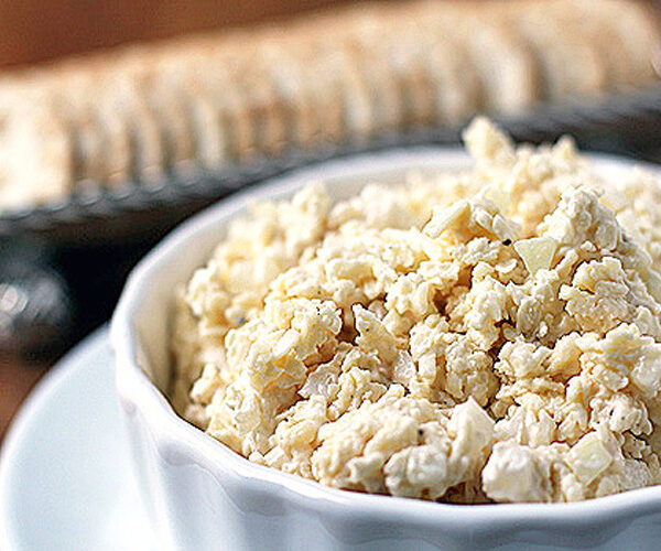 A white bowl filled with chunky homemade pimento cheese spread sits on a plate, with a stack of crackers blurred in the background.