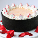 A round cake with a dark chocolate crumb crust, topped with white whipped cream and peppermint candies, sits on a white platter with red ribbon accents.