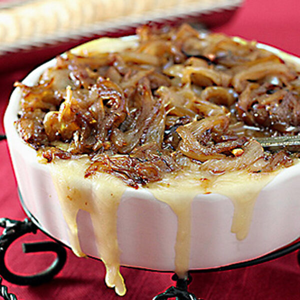 A white dish filled with hot, melted cheese topped with caramelized onions, with cheese dripping down the side. A spoon is in the dish, and crackers are visible in the blurred background on a red tablecloth.