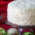 A round coconut-covered cake on a glass stand, set against a red background with a green ribbon and a silver ornament nearby.