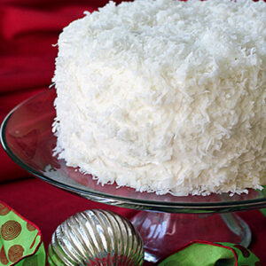 A round coconut-covered cake on a glass stand, set against a red background with a green ribbon and a silver ornament nearby.