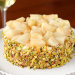 A round dessert topped with diced pears, coated on the sides with chopped pistachios, sits on a white plate. A glass of white wine is visible in the background.